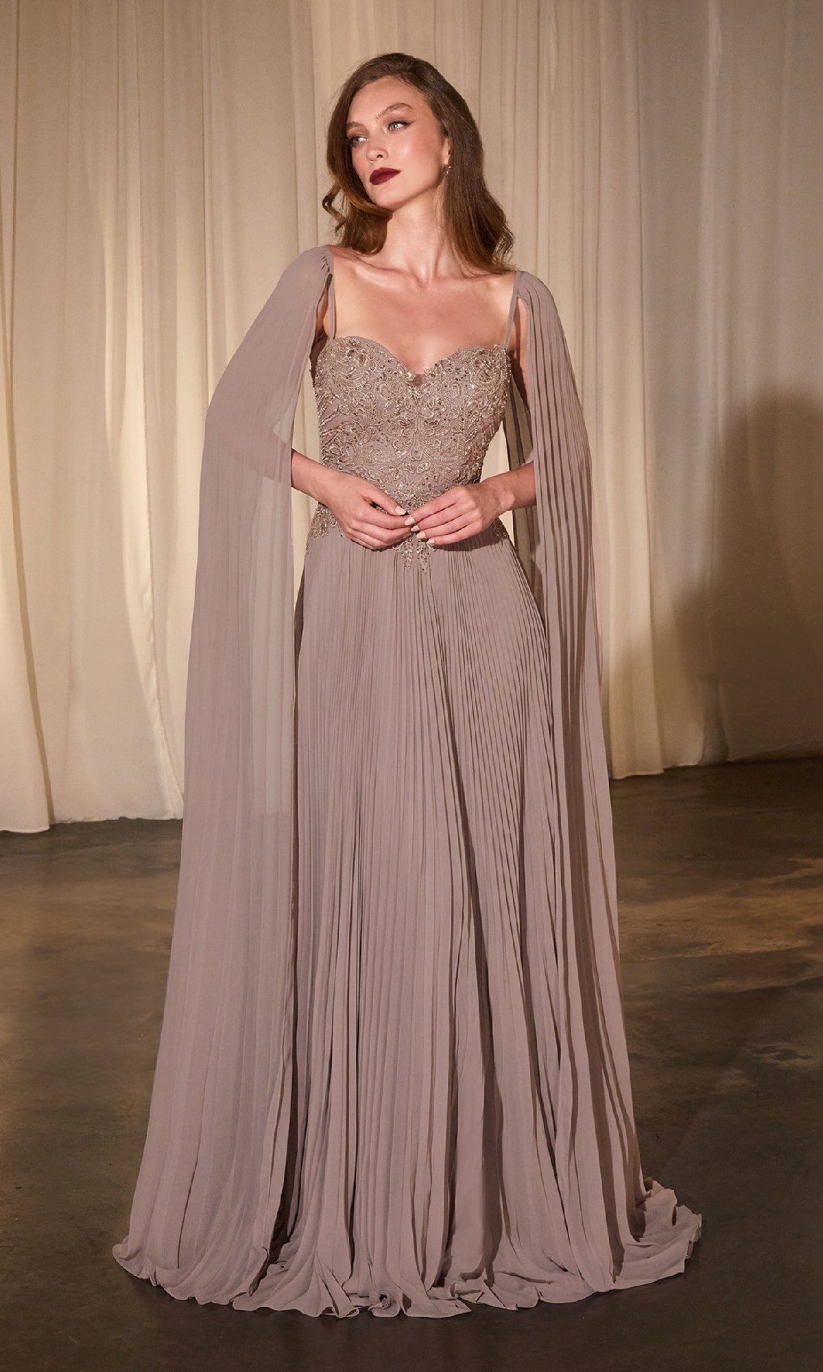 A woman in the Ladivine CJ937 Shoulder-Cape Long Prom Dress, featuring a taupe pleated skirt, beaded sweetheart bodice, and sheer cape sleeves, stands before beige curtains.